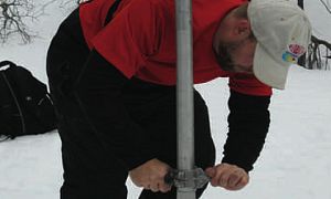 A man, wearing snow shoes, pushes a metal tube deep into the snow.  He is conducting a snow survey and helping measure Snow Water Equivalent (SWE).  